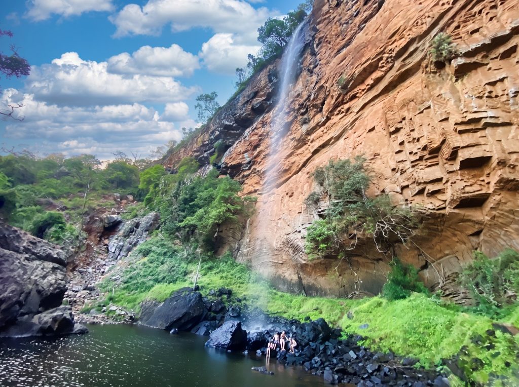 A tall waterfall dropping down a layered sandstone cliff into a green pool, with tiny figures at the water's edge